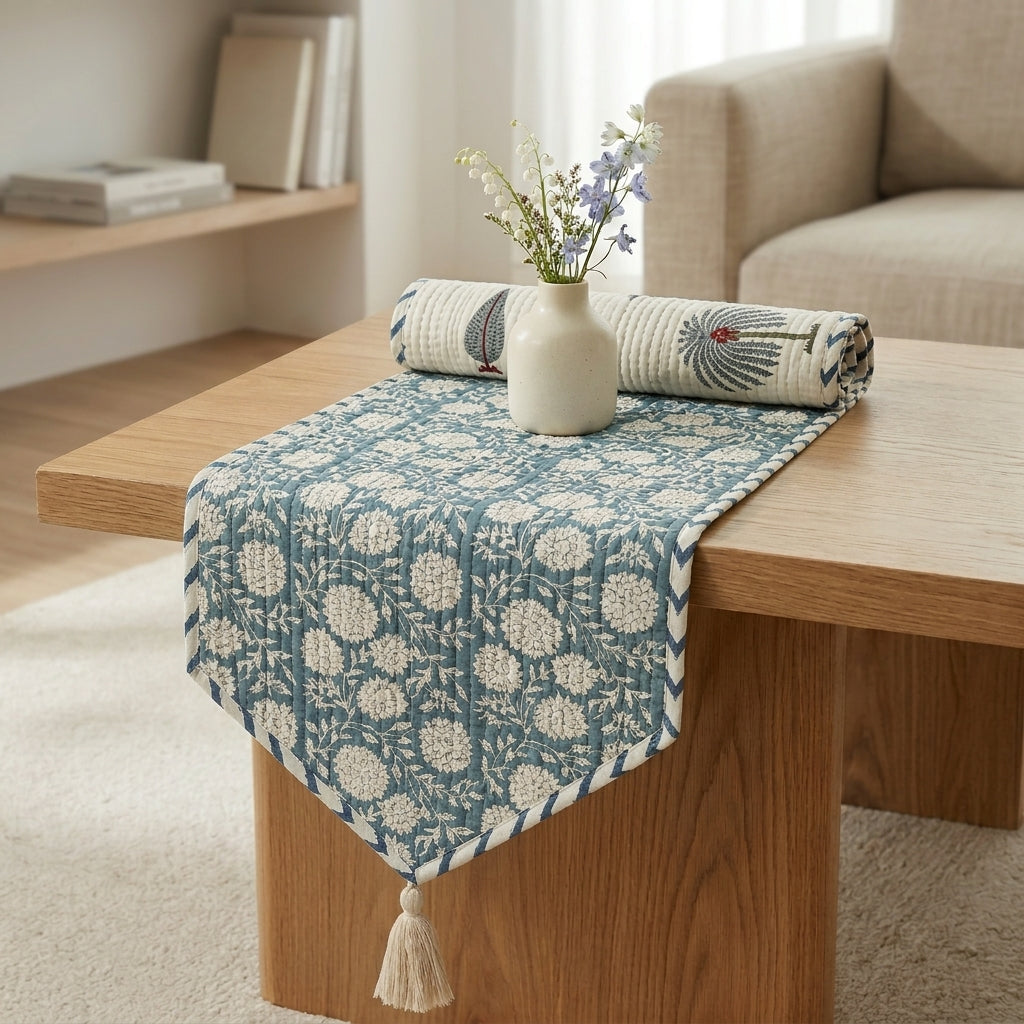 A lifestyle product photo of the quilted table runner from the reference image (with white floral damask print on dusty teal background), styled partially rolled up on a light-oak rectangular wooden table. A small cream ceramic vase with delicate wildflowers sits on top. Soft-focus bright living room background, warm natural daylight.