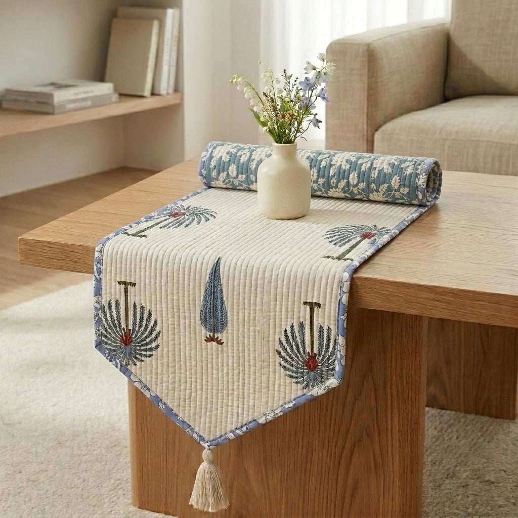 The same coffee table lifestyle scene, showing the reversible channel-quilted table runner. The unrolled front draping side displays the palm tree pattern with a periwinkle floral border, while the rolled-up portion resting on the table behind the ceramic vase displays its reverse side: a matte slate-blue or teal fabric featuring an intricate off-white floral damask pattern.